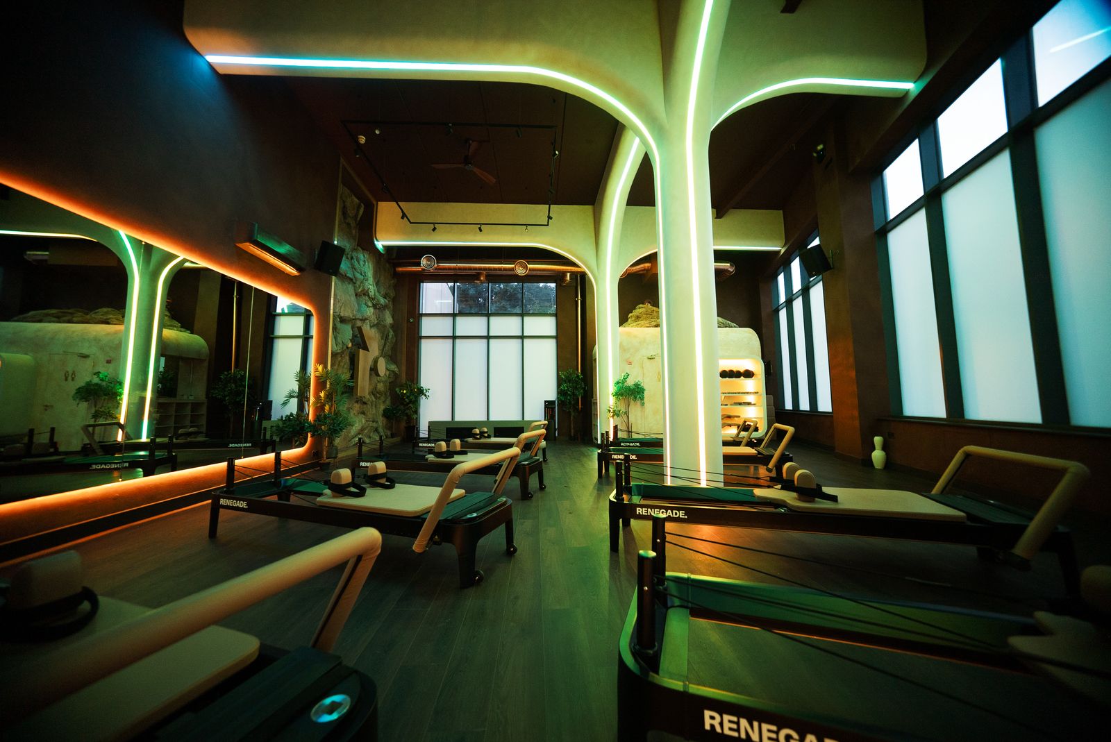 Wide view across the Renegade Reformer studio floor during a By Night session, green and amber neon arches overhead and Renegade branded reformer beds in the foreground